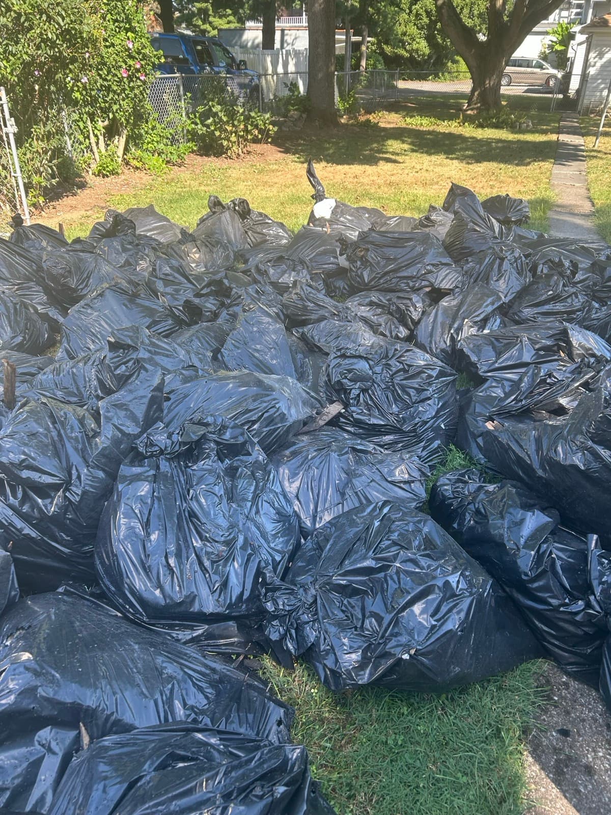 Row of filled black contractor bags lined along a chain-link fence on a grass strip — The Junk Lord