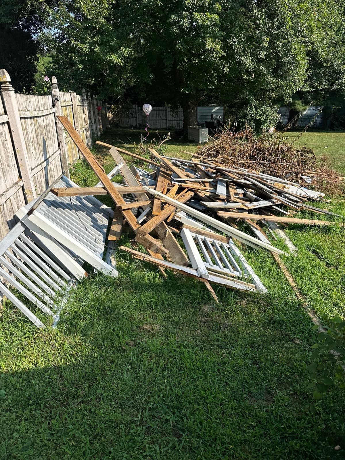 White porch railings, balusters, and broken framing piled on the lawn along a wood fence — The Junk Lord, York County PA