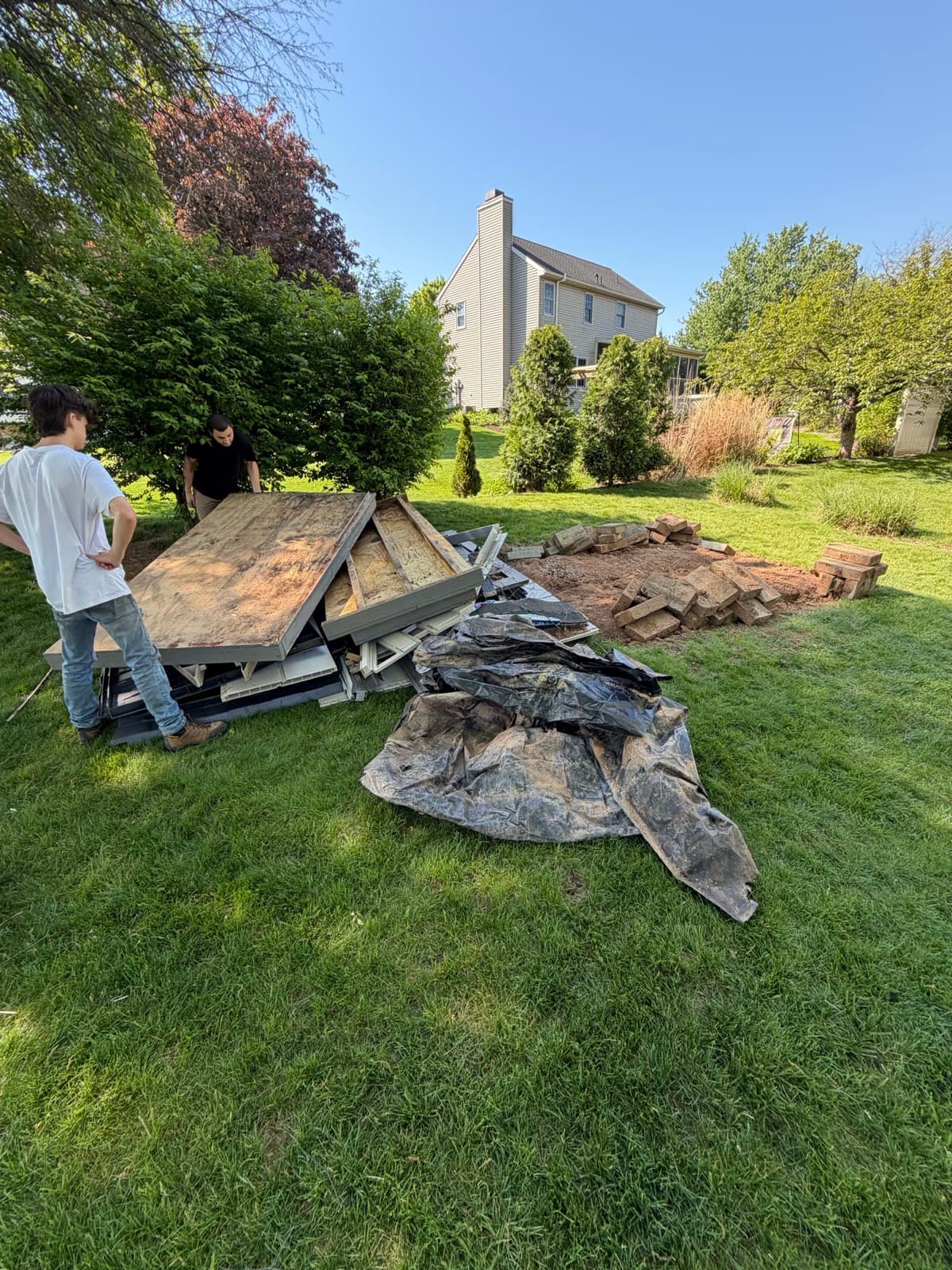 Two workers beside stacked plywood sheets and a tarp of demo debris on the lawn — The Junk Lord